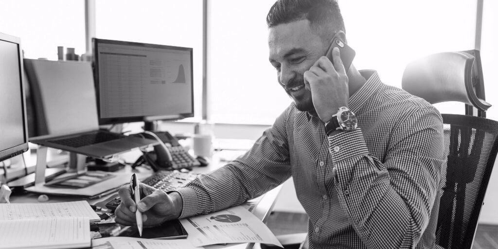 Man in collared dress shirt and watch smiles as he talks on the phone and holds a pen over papers featuring pie charts on a desk with multiple computer screens. 