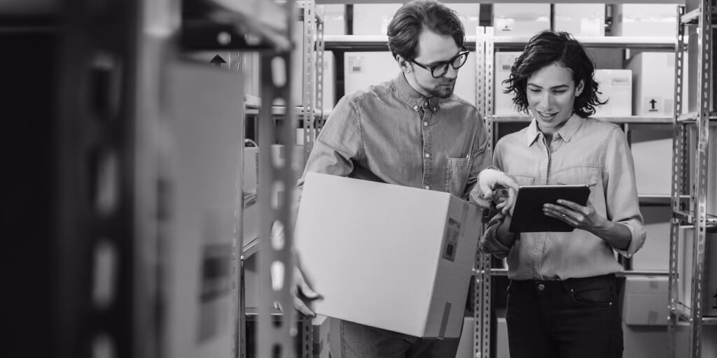 Two warehouse workers are seen discussing logistics in a storage area. The man on the left is holding a cardboard box and wearing glasses and a blue denim shirt. The woman on the right is holding a tablet, also dressed in a blue denim shirt. They appear to be reviewing inventory or shipment details. Shelves filled with boxes & packages are visible in the background, indicating an organized storage environment. The setting suggests collaboration & efficiency in a warehouse or distribution center.