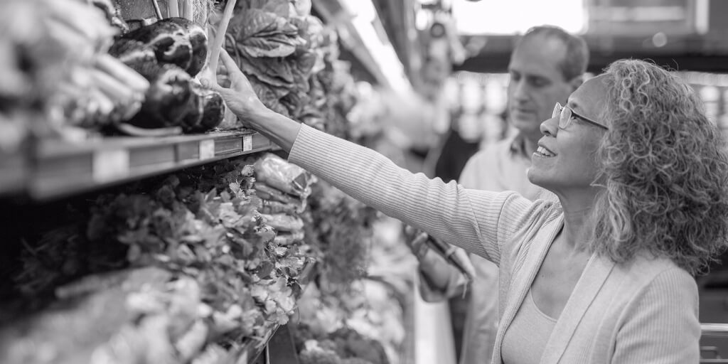 A smiling woman with curly gray hair and dark glasses reaches for a green vegetable among shelves of colorful produce at a grocery store.