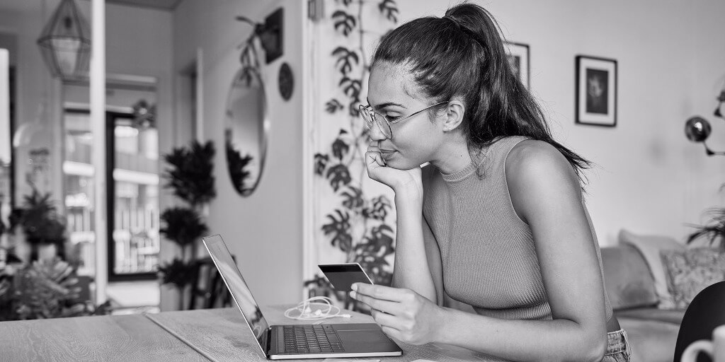 At a table in a modest living space, a young woman with long dark hair, wire-frame glasses, and an orange sleeveless shirt looks contemplatively at her laptop screen, a blue credit card in her hand.
