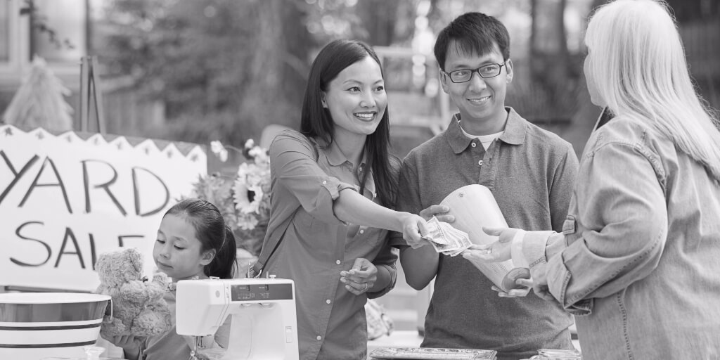 At an outdoor yard sale in front of tables laden with miscellaneous home supplies – including dishes and a sewing machine – a smiling, well-dressed couple buys a wooden vase from a white-haired woman in a denim jacket while their young daughter examines a teddy bear. 