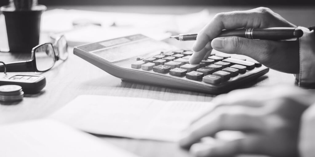 On a desk that holds scattered papers, a small cactus plant, eyeglasses, and a set of car keys, a man’s hand holds a red and gold fountain pen while typing on a calculator, the other hand resting on a slip of paper nearby.