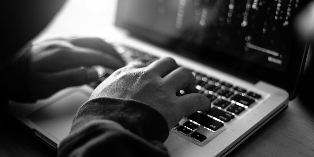 A man in a black hoodie hunches over the keyboard of a laptop that shows lines of binary code on the screen.