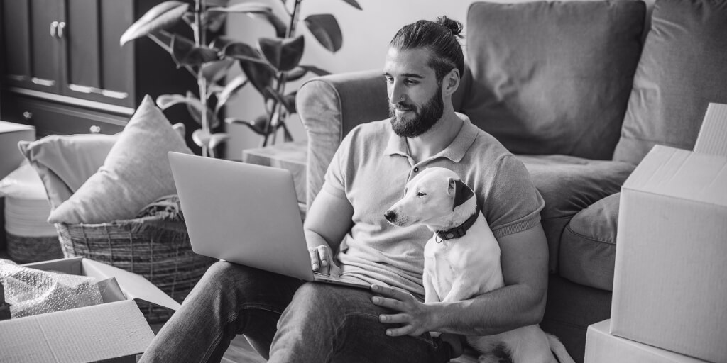 A smiling man in a tan t-shirt and blue jeans sits on the floor of a living room with a laptop in his lap and a small white dog nestled under his arm, with moving boxes stacked throughout the room around them.