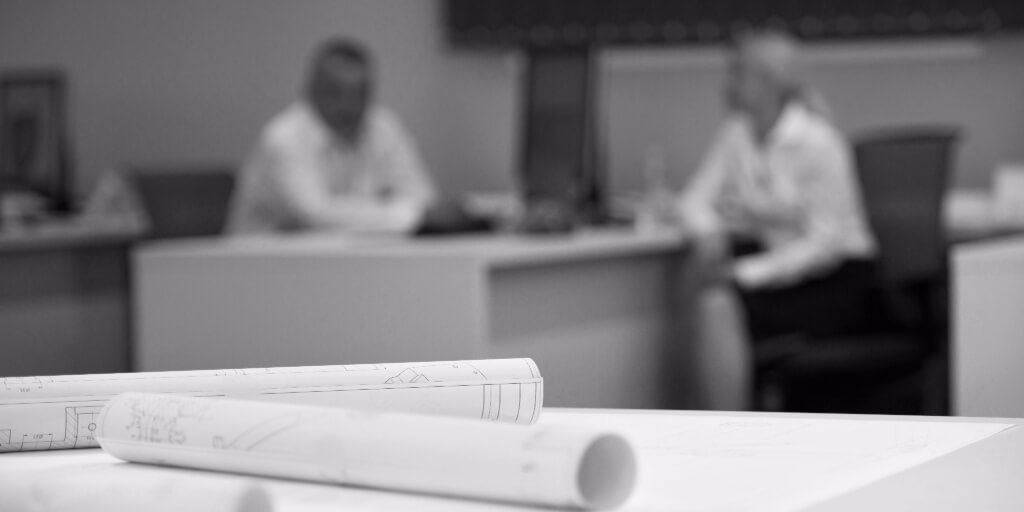 Close-up image of rolled blueprints with an out-of-focus of man and woman in business attire sitting at a desk in the background