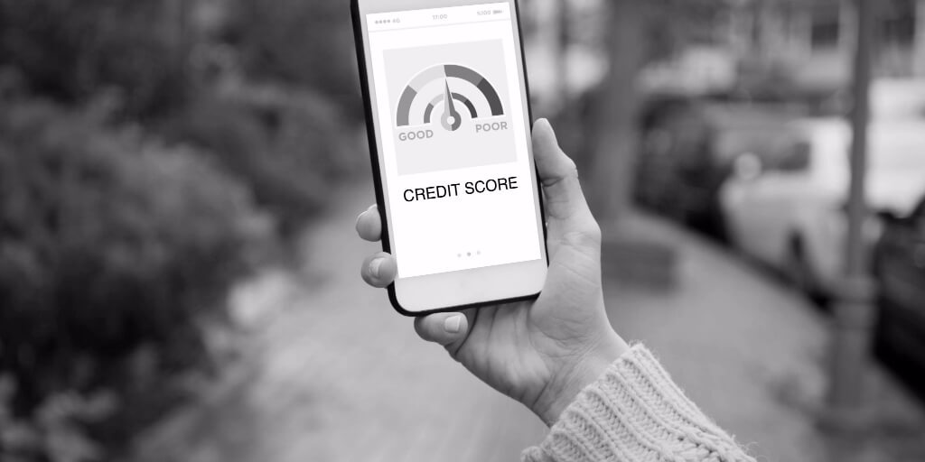 On a cloudy day and empty brick walkway, a woman’s hand holds up a cell phone whose screen displays a credit score meter ranging from good, represented by the color green, to poor, represented by the color red.