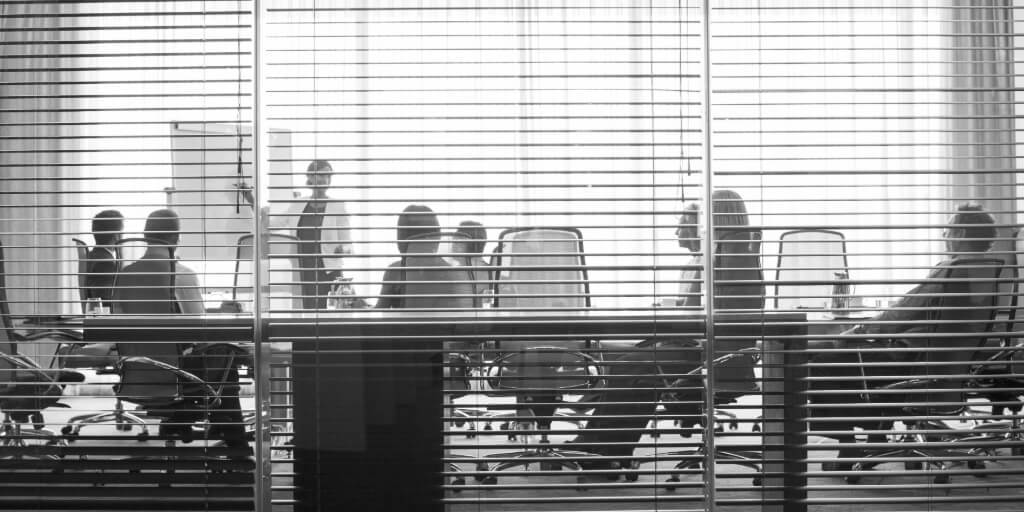 Through the slats of open blinds along a glass wall in an office, a woman in a black outfit with a white cardigan can be seen giving a presentation in a large conference room, as seven other professionals listen intently.