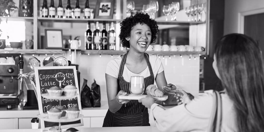 A joyous young woman wearing a black-and-white pinstriped apron hands a thick wooden plate with a cup of coffee and a croissant on it to a long-haired female customer. A variety of muffins sit on a stand between them in front of a small chalkboard that lists the prices for popular drinks like a cappuccino, latte, espresso, Americano, and mocha.