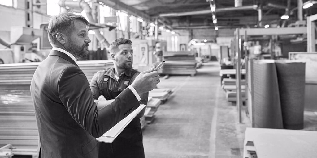 In an expansive warehouse full of lumber, a man with short gray hair in a blue suit holds a clipboard and points with a silver pen at something in the distance, while another man in blue overalls and a work shirt looks where he’s pointing.