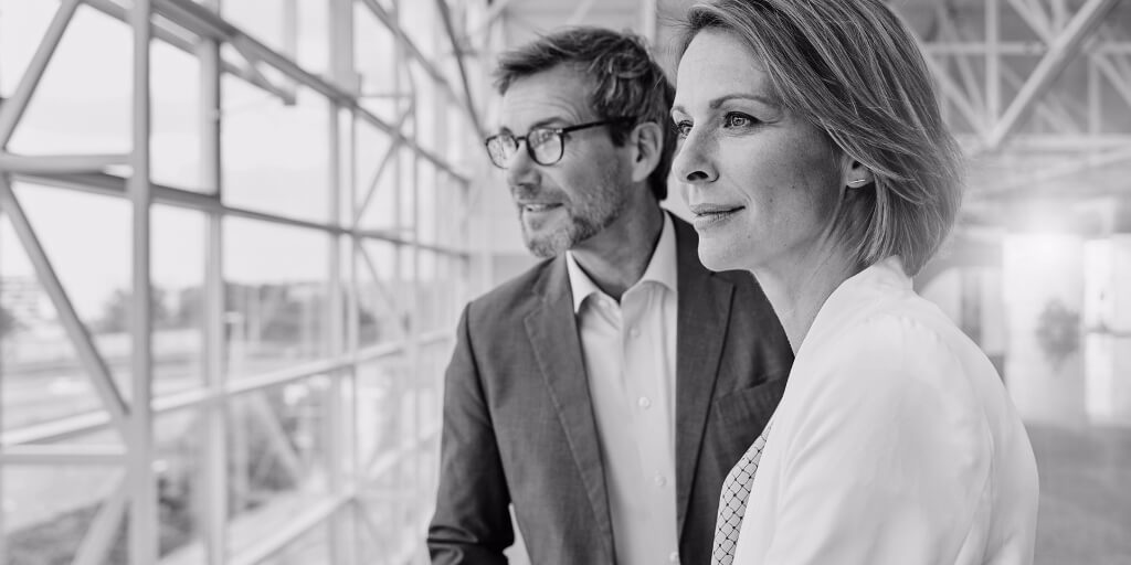 Woman in white blazer and man in glasses and gray blazer smile slightly as they look outside through a wall of windows