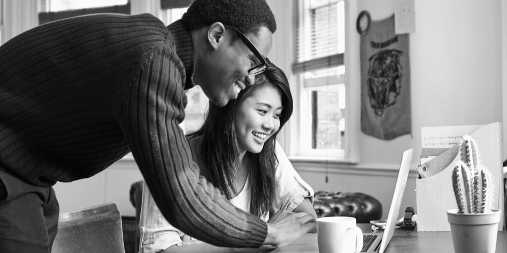 Sitting at a black wooden desk in front of three windows in a modest apartment, a dark-haired young woman in a white shirt smiles widely as a man in dark glasses, a dark blue sweater, and red pants leans down to use the silver laptop open on the desk in front of them.
