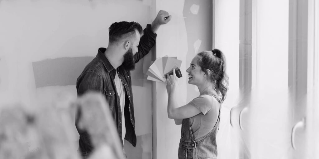 A young couple is standing in a partially renovated room, discussing paint colors while one person holds a fan of color swatches