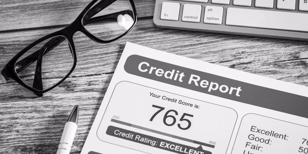 A close-up shot of a desk focuses on a printed credit report with an excellent credit rating and a credit score of 765. A white pen, black glasses, and gray and white keyboard rest on the desk around the credit report.
