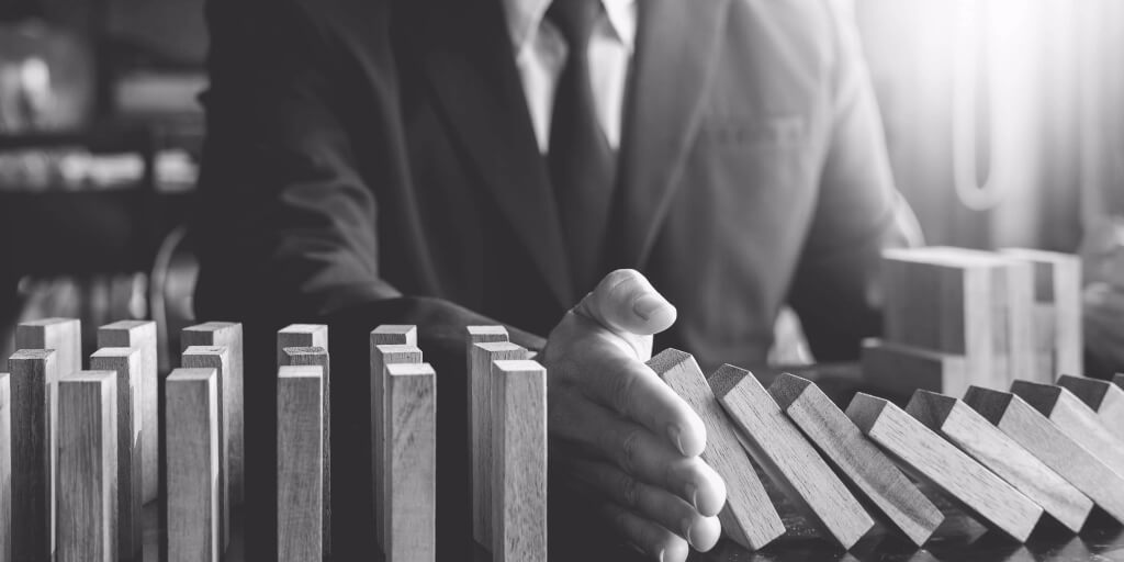 Close-up image of a man in a business suit trying to stop a row of wood blocks from falling over like dominoes on his desk